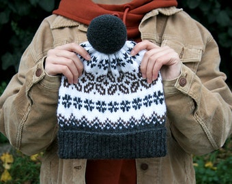 May include: A white, brown, and black knit beanie with a pom pom on top. The beanie has a traditional Scandinavian pattern.
