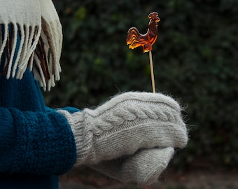 Può includere: Una persona che indossa un maglione blu e una sciarpa bianca tiene un lecca-lecca marrone a forma di gallo su un bastoncino. La persona indossa guanti di lana grigi.