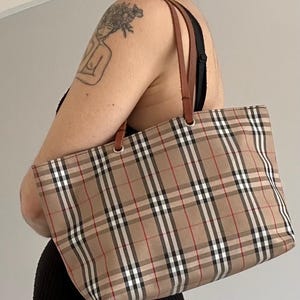 May include: A beige tote bag featuring a traditional plaid design in black, white, and red. It has brown leather handles and a rectangular form. The bag is held by a person.