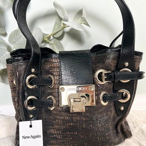 Peut inclure: Sac à main Jimmy Choo marron et noir avec un motif texturé. Le sac présente des poignées, des accents et une quincaillerie noirs. Un fermoir et des anneaux argentés ornent le devant. Une étiquette blanche avec "New Again" et "Vinted" est suspendue à la poignée.