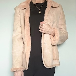 May include: A light tan suede jacket with a shearling collar and trim. The jacket has a button closure and two front pockets. The lining is a light pink color. The jacket is worn over a black outfit.