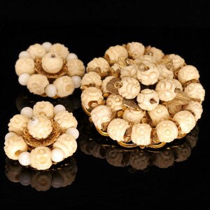 May include: A set of three vintage brooches made of carved ivory-coloured beads and gold-toned metal. The brooches are in the shape of flowers with a central bead and a cluster of smaller beads around it.