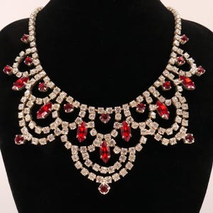 Può includere: Una collana a pettorina caratterizzata da una serie di strass trasparenti sfaccettati disposti a festone. Il design incorpora diverse pietre rosse a taglio marquise e rotondo incastonate nella struttura in metallo color argento.