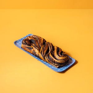 May include: A rectangular blue and white patterned ceramic serving tray holding a golden-brown braided pastry with dark chocolate or poppy seed swirls. The pastry has a layered, twisted texture and is positioned centrally on the decorative dish.