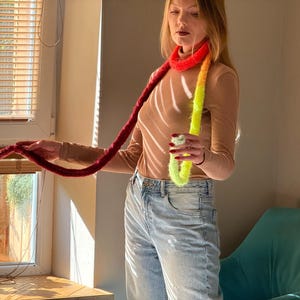 May include: A long, colorful scarf with a gradient of colors from red to yellow. The scarf is made of a soft, fuzzy material. The person is wearing a tan long-sleeve shirt and blue jeans. The scarf is wrapped around the neck.