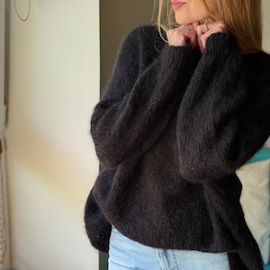 May include: A dark charcoal-colored, oversized sweater with long sleeves. The sweater has a soft, fuzzy texture and a relaxed fit. The image shows the sweater from the shoulders down, with a glimpse of blue jeans.