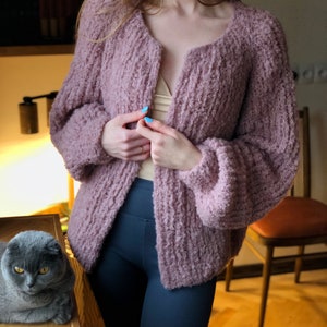 Puede incluir: Un cárdigan suave y peludo de color rosa con cuello en V y mangas largas. El cárdigan está abierto y la persona que lo lleva está sujetando los bordes del cárdigan.