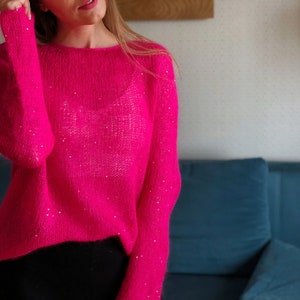 Puede incluir: Un suéter de color rosa brillante con una textura brillante. El suéter tiene mangas largas y un cuello redondo.