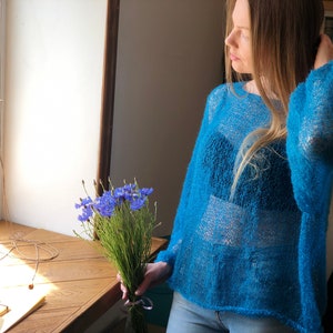 May include: A woman wearing a light blue, loose-fitting, knitted sweater with a sheer texture. She is standing near a window with a bouquet of blue flowers in her hand.