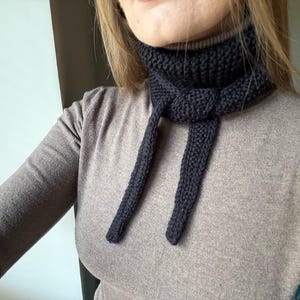 May include: A dark navy blue knitted scarf with long ties, tied in a knot at the neck. The scarf is made of a thick, textured yarn and is worn over a light brown long-sleeved shirt. The scarf is a winter accessory.