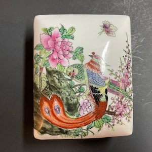 May include: A white porcelain box with a square shape and a floral design. The box features two birds, one with a long tail, and a butterfly. The birds are painted in shades of red, orange, green, blue, and black. The flowers are painted in shades of pink, purple, and green.