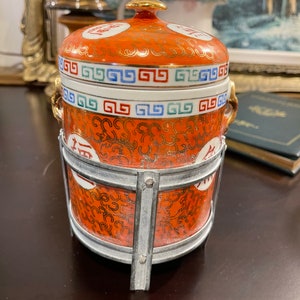Peut inclure: Un pot en céramique rouge et or avec un couvercle et un support en métal. Le pot présente un motif décoratif de motifs tourbillonnants et de caractères chinois. Le couvercle a un bouton doré et une bande blanche avec un motif répétitif.
