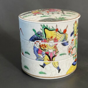 May include: A white ceramic tiered container with a lid, decorated with colourful hand-painted figures and floral designs. The container has a crackled glaze finish.