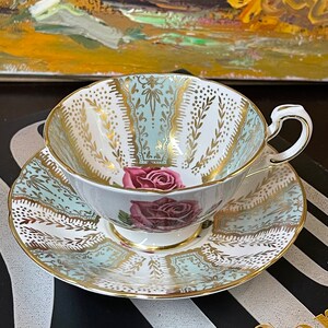 May include: A delicate teacup and saucer set with a floral design. The cup and saucer are white with a light blue and gold pattern. The floral design features a pink rose with green leaves.