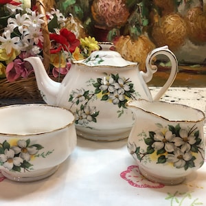 May include: A white porcelain tea set featuring a teapot, creamer, and sugar bowl. Each piece is decorated with a floral pattern of white flowers, green leaves, and yellow accents, with gold trim. The set is arranged on a white tablecloth.