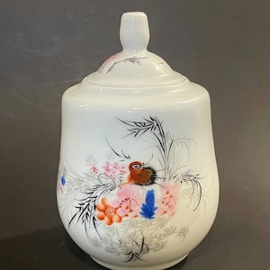 May include: White ceramic jar with a lid, decorated with a painted bird, flowers, and bamboo. The bird has brown, orange, and black feathers. The flowers are pink, orange, and blue. The jar has a rounded shape.