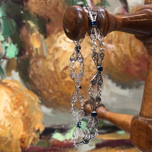 May include: A silver filigree bracelet with blue enamel accents. The bracelet is displayed on a wooden stand.
