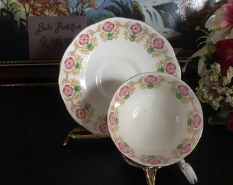 May include: A white teacup and saucer set with a floral pattern in shades of pink, green, and gold. The set is on a gold metal stand.