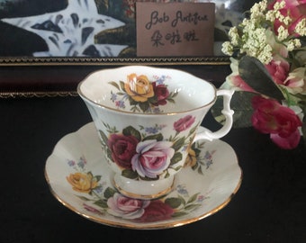 May include: A white teacup and saucer with a floral pattern featuring pink, red, and yellow roses. The cup has a gold rim and the saucer has a scalloped edge.