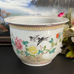 Peut inclure: Un pot en céramique blanche avec un motif floral composé de fleurs roses et jaunes, de feuilles vertes et de deux oiseaux noirs. Le pot est orné d'une bordure dorée et violette autour du haut.