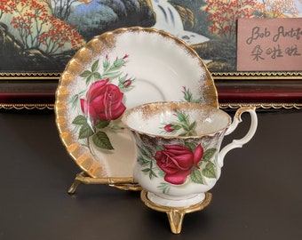 May include: A white teacup and saucer with a gold rim and a red rose design. The teacup and saucer are on a gold metal stand. The text "Bob Antique" and Chinese characters are visible in the background.