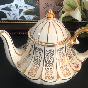 May include: A white teapot with a gold lid and intricate gold designs. The teapot has a rounded body and a curved spout.