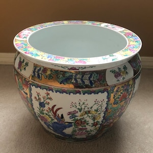 May include: A large, white porcelain planter with a round, wide base and a flared rim. The planter is decorated with a colourful floral pattern in shades of pink, blue, green, yellow, and gold. The pattern includes flowers, butterflies, and birds.