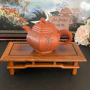 Peut inclure: Une théière en argile rouge avec des caractères chinois et un motif de paysage repose sur un support en bois. La théière a un couvercle avec un petit bouton sur le dessus. Le support a quatre pieds et un dessus rectangulaire.
