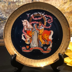 May include: A decorative porcelain plate with a gold rim and a blue background. The plate features a painting of a knight on horseback, holding a woman's hand. The knight is wearing a red and gold suit of armor, and the woman is wearing a white dress. The scene is surrounded by a garland of pink flowers and white doves.