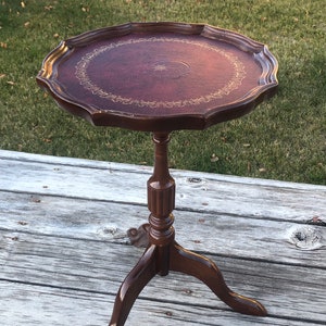 May include: A dark brown, antique-style side table with a round, scalloped top. The tabletop features a burgundy leather insert with a gold decorative border. The table stands 71 cm tall and has a tripod base.