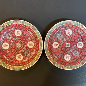 May include: Two red and white ceramic plates with a decorative design of stylised flowers and Chinese characters. The plates have a gold trim and a green border.