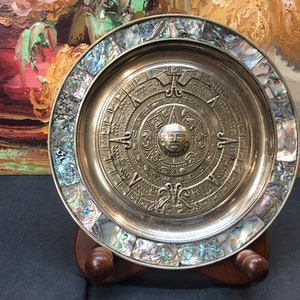 May include: A silver plate featuring a decorative design of the Aztec calendar, surrounded by a border of iridescent abalone shell. The plate is displayed on a wooden stand.