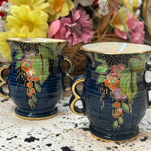 May include: Two small blue ceramic vases with gold trim and floral designs. The vases have two handles each and are sitting on a white lace tablecloth.