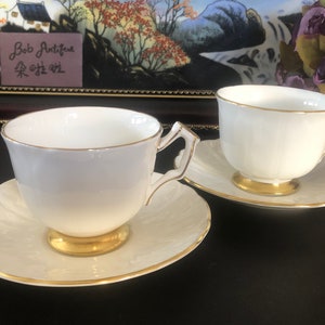 May include: Two white teacups and saucers with gold trim. The cups have a delicate, fluted design and are set on matching saucers. The saucers have a scalloped edge and a subtle, embossed pattern.