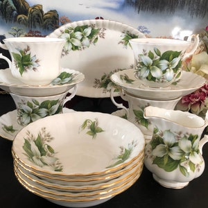 May include: A set of white bone china teacups and saucers with a floral pattern of white flowers and green leaves. The set includes a creamer and a serving platter. The set has a gold trim.