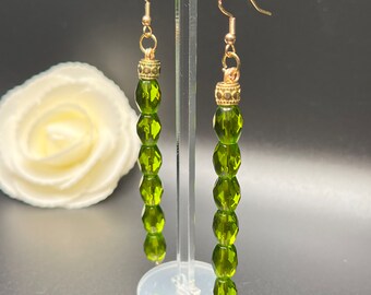 Puede incluir: Un par de aretes largos de cuentas de vidrio verde con detalles dorados. Los aretes están colgados de un soporte de plástico transparente.