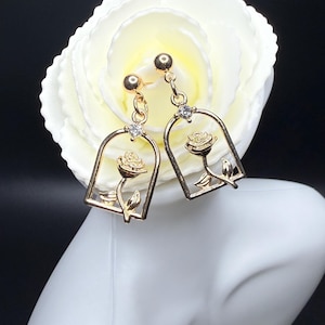 May include: Gold-toned dangle earrings featuring a rose design within a bell-shaped frame. Each earring has a small, clear gemstone and a post. The earrings are displayed against a white flower and a black background.