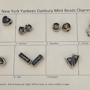 May include: A collection of New York Yankees Danbury Mint beads charms. The charms include heart, helmet, baseball, jersey, glove, bar, and pennant designs. Each charm features the Yankees logo in navy blue and white.