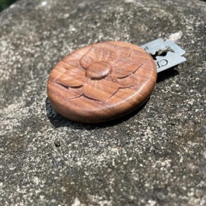 Puede incluir: Una caja redonda de madera con un diseño de flor tallado en la parte superior. Una sola cuchilla de afeitar está parcialmente insertada en la caja.