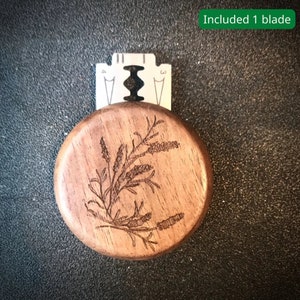 Könnte beinhalten: Ein runder Rasierergriff aus Holz mit einem detaillierten, eingravierten Blumendesign. Der Griff besteht aus hellbraunem Holz und enthält eine silberne Rasierklinge. Der Text "Included 1 blade" befindet sich in einem grünen Rechteck.