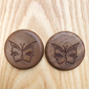 May include: Two round wooden coasters with a butterfly design carved into the surface. The coasters are dark brown and have a natural wood grain.