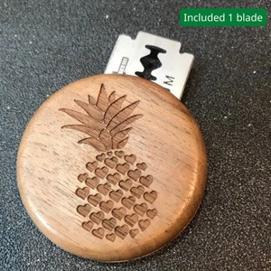 May include: A wooden, round, engraved box with a pineapple design. The pineapple is made up of hearts. The box is open and a single razor blade is visible inside. The text "Included 1 blade" is visible in the upper right corner of the image.