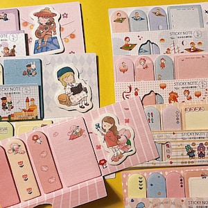 Può includere: Assortimento di note adesive colorate con varie illustrazioni di personaggi dei cartoni animati, tra cui una ragazza con un gatto, una ragazza con un libro, una ragazza con un fiore e una ragazza con una barca. Le note adesive sono etichettate "STICKY NOTE" e hanno il numero "6" su di esse.