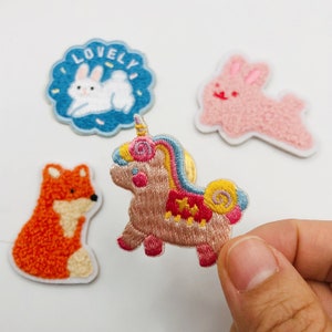 Peut inclure: Un ensemble de cinq patchs brodés thermocollants représentant un lapin rose, un lapin bleu avec le mot "LOVELY", un renard et une licorne.