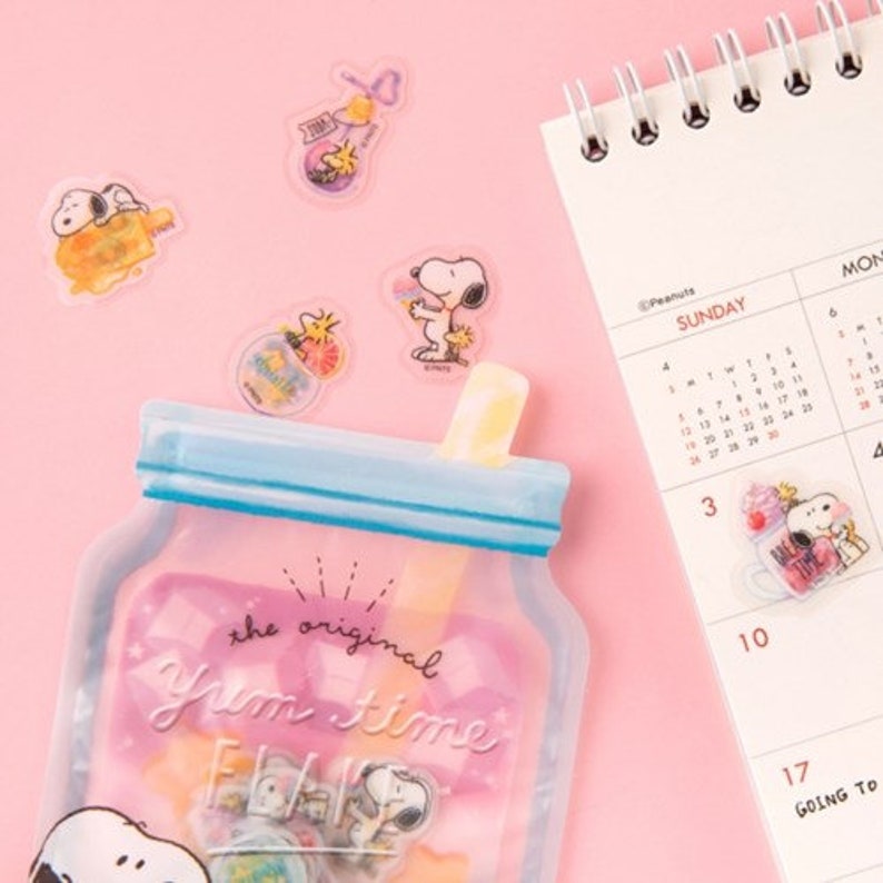 May include: A clear plastic bag with a blue zipper and a straw on top. The bag has a pink background with the text "the original Yum Time" printed on it. There are several clear stickers with cartoon characters on the bag. The stickers feature a cartoon dog with a red nose and a yellow collar.