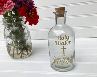 Puede incluir: Una botella de vidrio transparente con tapón de corcho, con letras doradas que dicen Holy Water, un símbolo de cruz dorada y el texto Eva Marie 10/23/22. Al lado hay un frasco de vidrio lleno de flores de zinnia rojas, rosas y moradas.