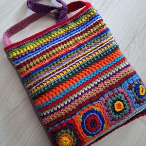 Może przedstawiać: Kolorowa, dziana torba na ramię z fioletowym paskiem. Torba wykonana jest r&oacute;żnymi ściegami i kolorami, w tym czerwonym, pomarańczowym, ż&oacute;łtym, zielonym, niebieskim i fioletowym. Torba ma kwadratowy kształt i jest ozdobiona kilkoma dzianymi kwiatami.