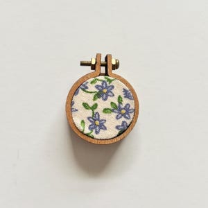 May include: A small wooden embroidery hoop with a white fabric background and a floral pattern in shades of purple and green.
