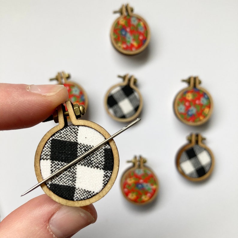 May include: A set of six small embroidery hoops with different fabric patterns. The hoops are made of wood and have a metal screw closure. The fabrics include black and white gingham, red and white floral, and blue and white floral.