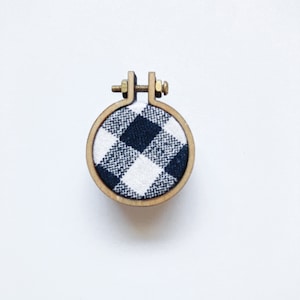 May include: A small wooden embroidery hoop with a black and white checked fabric inside. The hoop is approximately 5 cm in diameter.